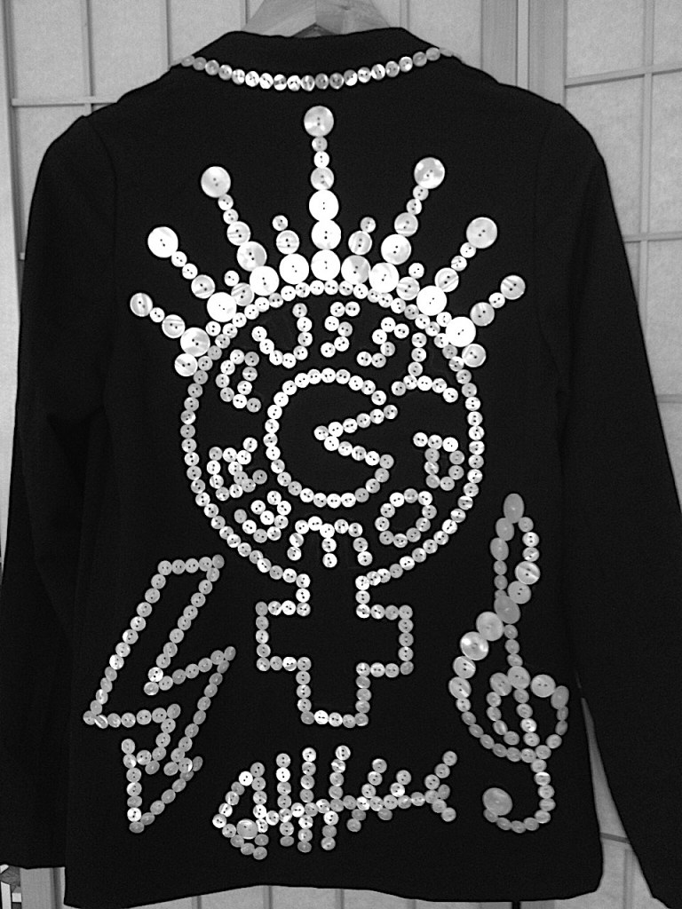 A design featuring a crown, women’s symbol, Pacman, lightning bolt, fish skeleton, and treble clef, all made of pearl buttons sewn onto black fabric